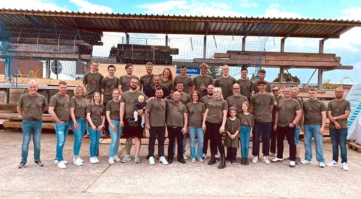 Eine Gruppe von Menschen steht draußen zusammen, alle tragen olivgrüne T-Shirts und lächeln in die Kamera. Im Hintergrund ist ein Zaun und ein Unterstand zu sehen.