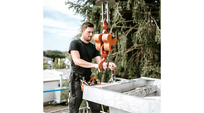 Ein Mann bedient einen großen orangefarbenen Kranhaken und befestigt ihn an einem Metallträger im Freien.