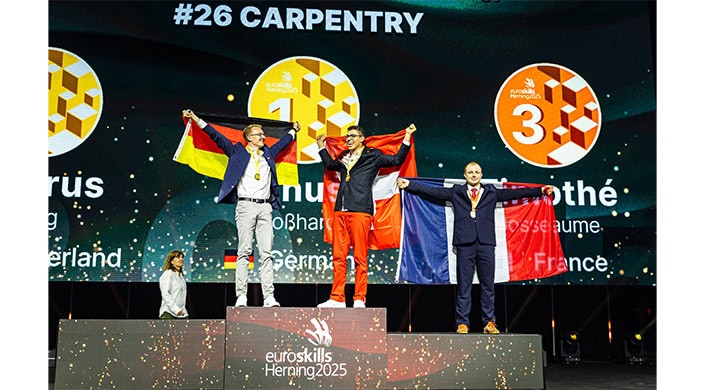 Ein Mann steht jubelnd auf einem Siegerpodest, trägt eine Medaille und hält eine rot-weiße Flagge, umgeben von anderen Flaggen.
