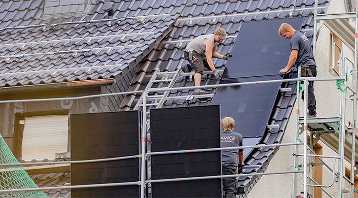 Ein Arbeiter befindet sich auf einem Dach mit dunklen Ziegeln, steht auf einer Leiter und führt Dacharbeiten durch. Baugerüst im Vordergrund.