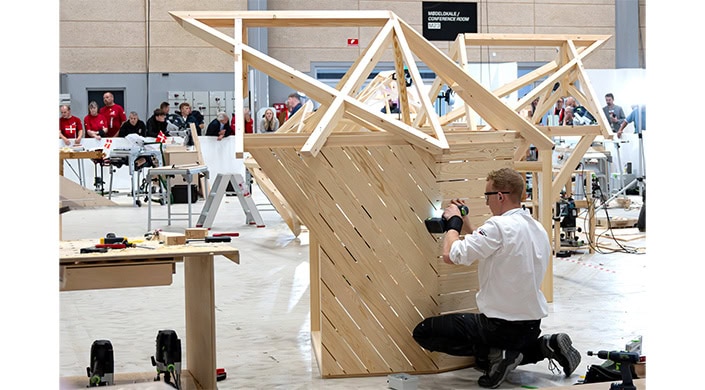 Holzkonstruktion mit diagonalen Latten und geneigten Trägern, vermutlich Teil eines Dach- oder Architekturmodells, im Innenraum mit einigen Menschen im Hintergrund.