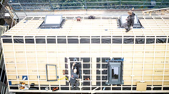 Arbeiter auf einem Dach montieren Holzbalken und schwarze Abdeckfolie, Bauarbeiten im Freien, Schutzmaßnahmen erkennbar.