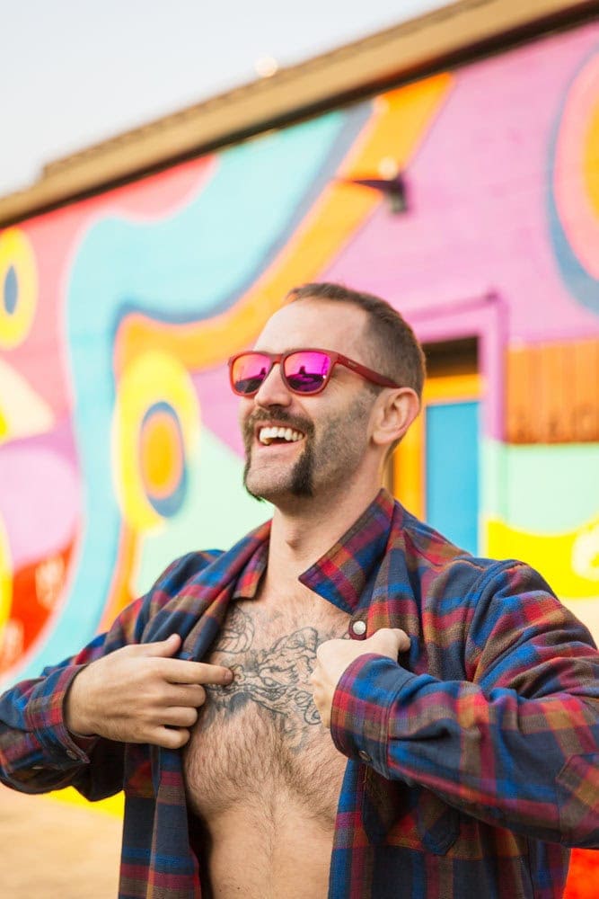 A man with a handlebar mustache and mirrored sunglasses is smiling and opening his plaid shirt to reveal chest tattoos. A colorful mural is in the background.