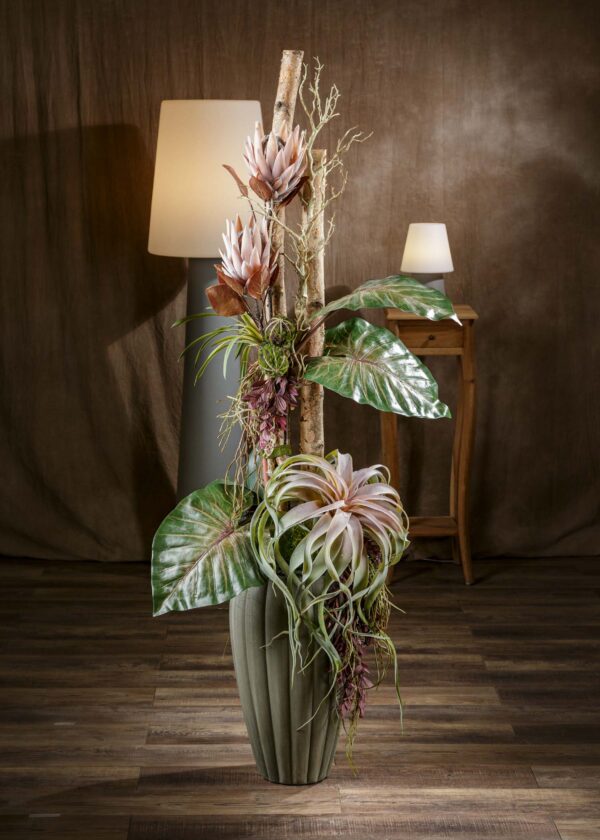 Vista frontale di una composizione floreale particolare con fiori artificiali e tronchi naturali di betulla. I fiori, dai colori tenui e dalle forme delicate, sono disposti con armonia tra le foglie verdi e i tronchi di betulla, creando un contrasto armonioso e raffinato. La composizione è collocata in un vaso di ceramica verde di design che completa l'eleganza dell'insieme. - Composizione floreale particolare, fiori artificiali, tronchi di betulla, vaso di ceramica verde.