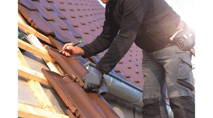 Ein Arbeiter mit Handschuhen befestigt eine Dachrinne an einem Hausdach mit roten Ziegeln.