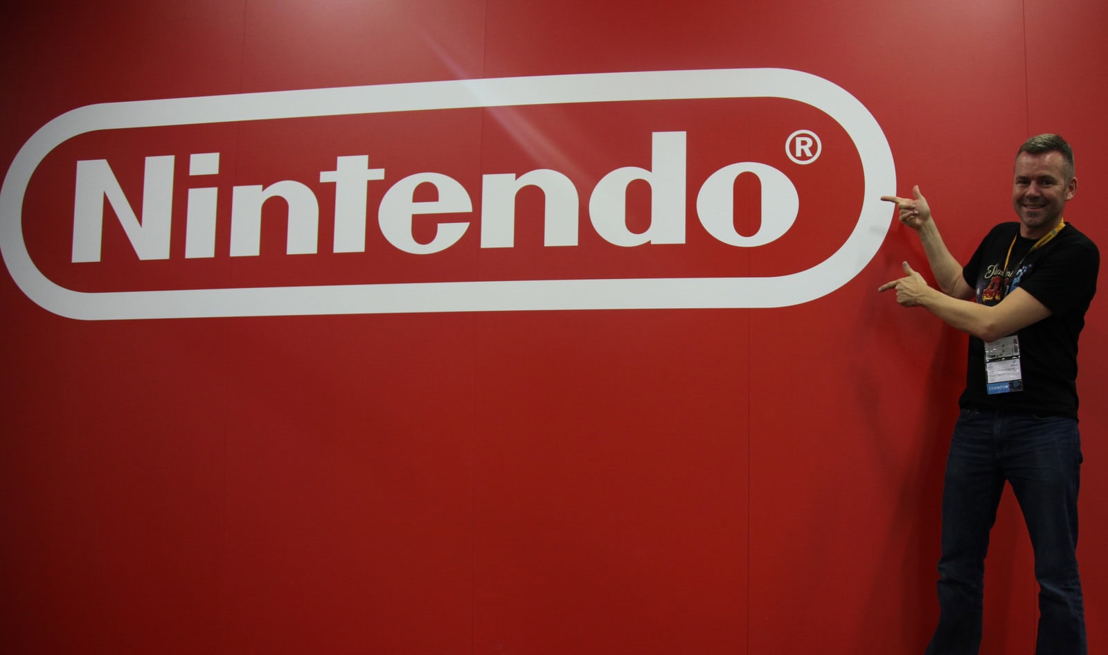Nintendo logo géant sur un mur rouge avec un homme souriant pointant le logo, lors d'une conférence ou événement gaming.