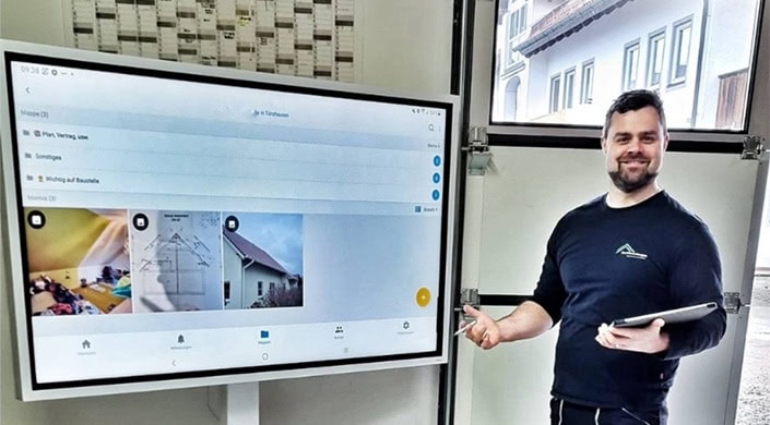 Ein großer Monitor zeigt eine Benutzeroberfläche mit Suchleiste, Symbolen und Schaltflächen, montiert an einer weißen Wand in einem modernen Raum.