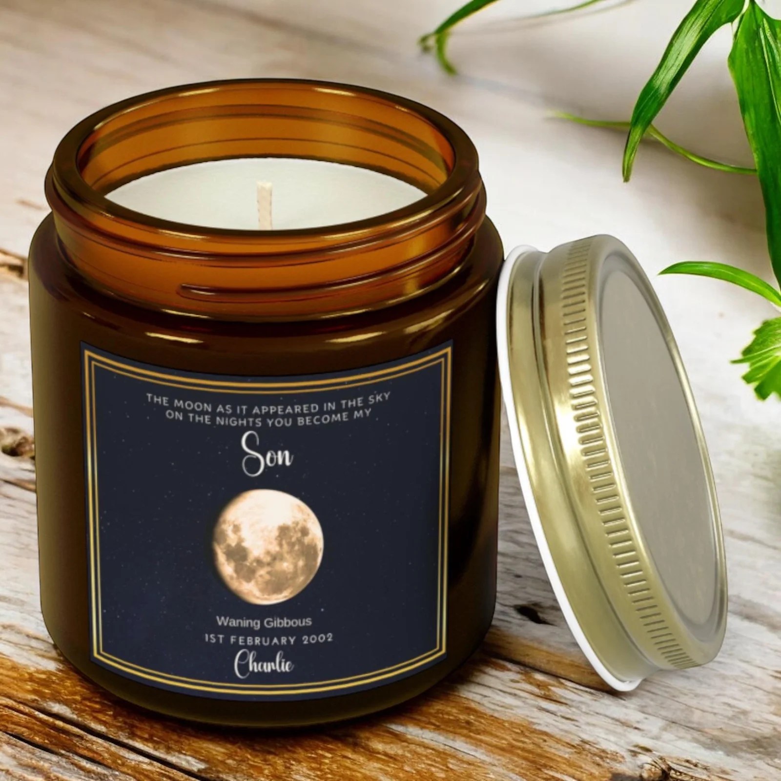 Scorpio Season 6 A brown glass candle jar with a silver lid on a wooden surface. The label shows a moon phase, text about the night a son was born, and the date 1st February 2002. Green leaves are visible in the background.