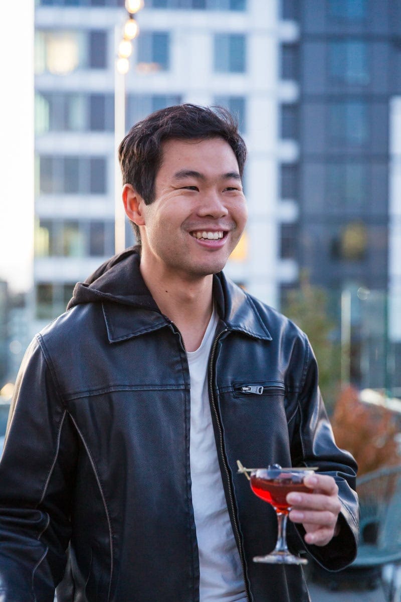 Person in a black jacket smiles while holding a cocktail on a rooftop with buildings in the background.