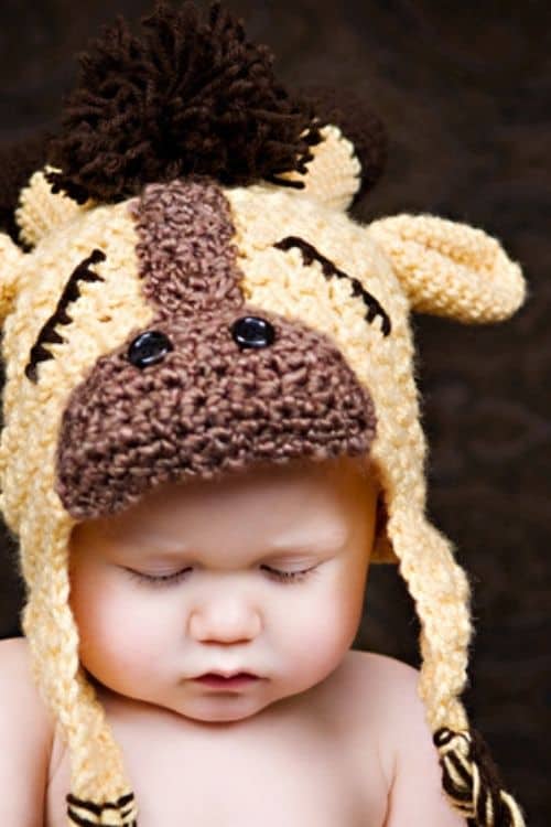 Baby sleeping, wearing a yellow and brown giraffe-themed crochet hat with ears, mane, and tassels on a dark background. Free crochet pattern