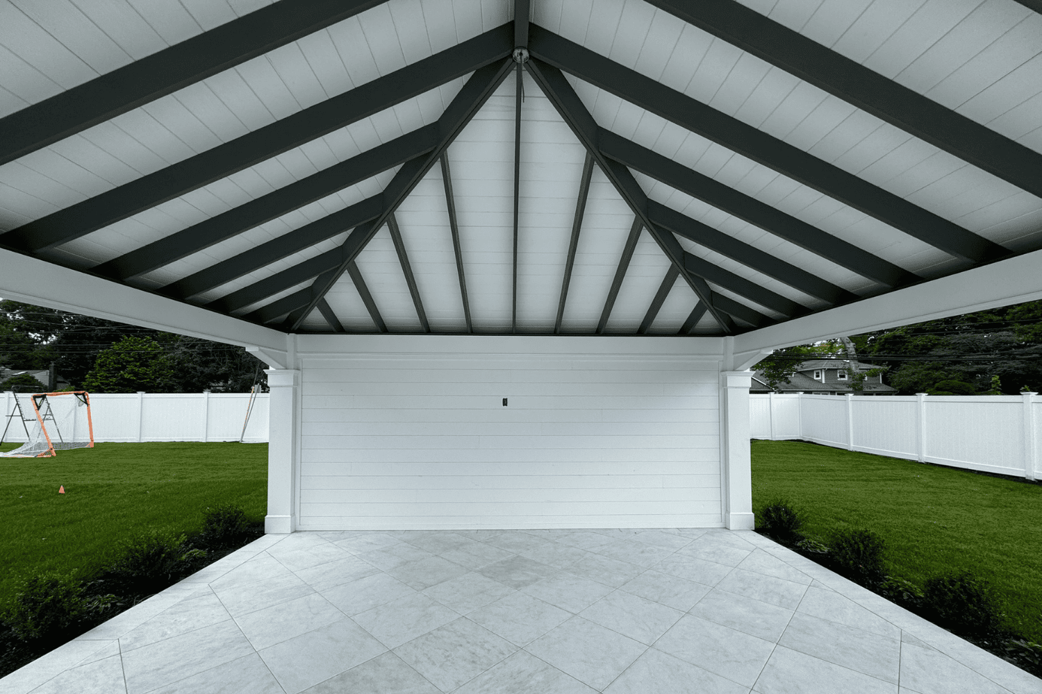 Covered patio with a white shiplap wall, gray exposed beams, and tiled flooring—perfect for relaxing or entertaining—overlooks a fenced backyard with grass, a swing set, and space ideal for pergolas or pavilions.