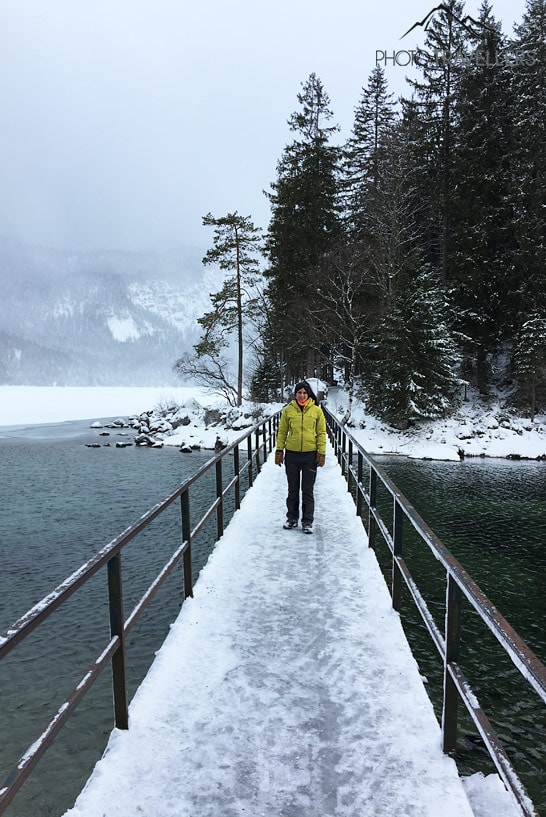 Reisebloggerin Biggi Bauer steht im Winter mit einer gelben Jacke auf der vereisten Brücke am Eibsee