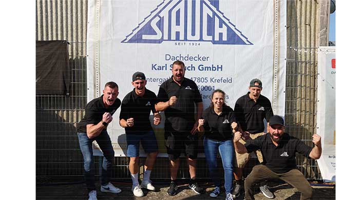 Drei Menschen in schwarzer Arbeitskleidung stehen vor einem Firmenplakat mit der Aufschrift „Stauch Dachdecker Karl Stauch GmbH“ und lächeln in die Kamera.