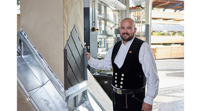 Ein Mann in traditioneller Handwerkskleidung arbeitet in einer modernen Werkstatt und hält ein Bauelement fest.