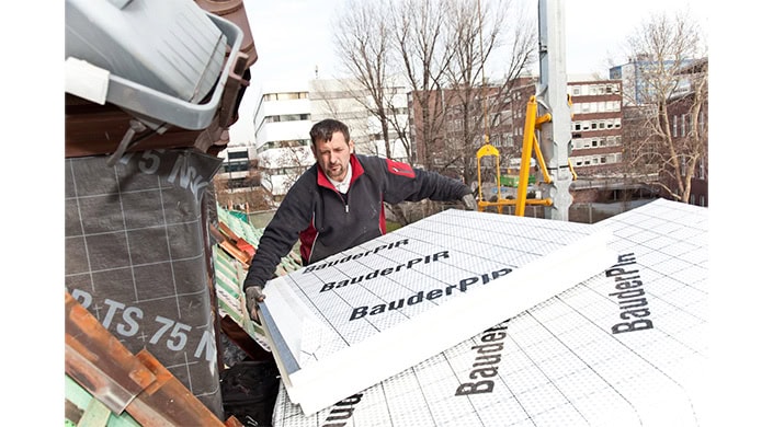 Ein Mann installiert weiße Dämmplatten mit der Aufschrift „BauderPIR“ im Freien, Bäume und Gebäude im Hintergrund.