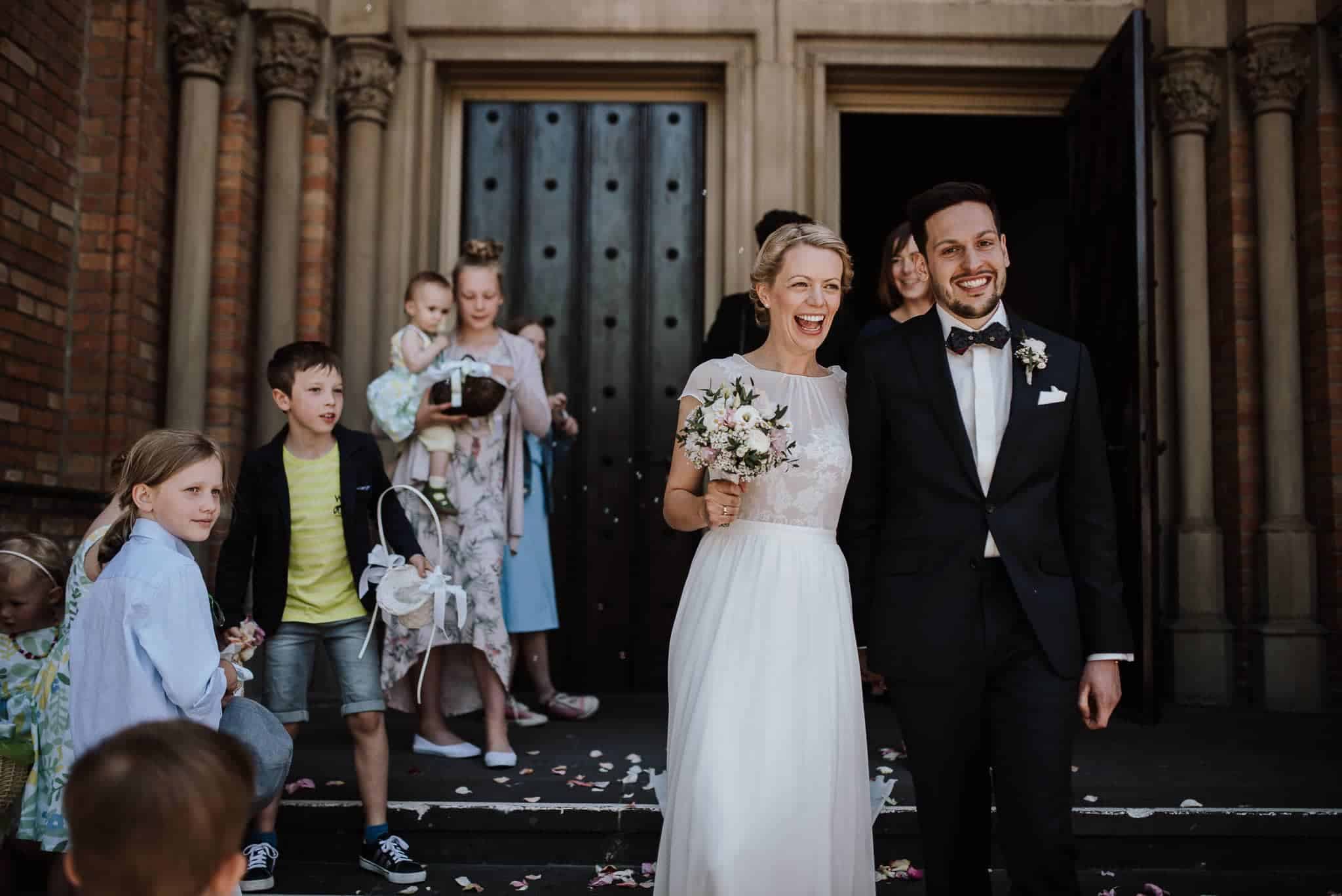 Frischvermählte auf Gut Hohenholz gehen Hand in Hand hinaus, Gäste und Kinder werfen Blütenblätter – Hochzeitsfotografin Köln.