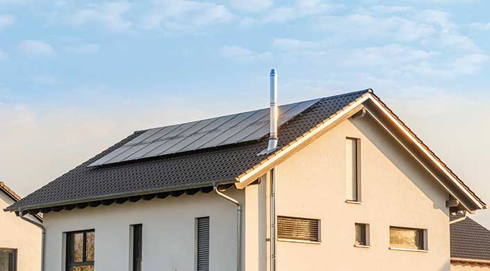 Dach eines Hauses mit Solarpaneelen und einem Schornstein bei Tageslicht, blauer Himmel im Hintergrund.