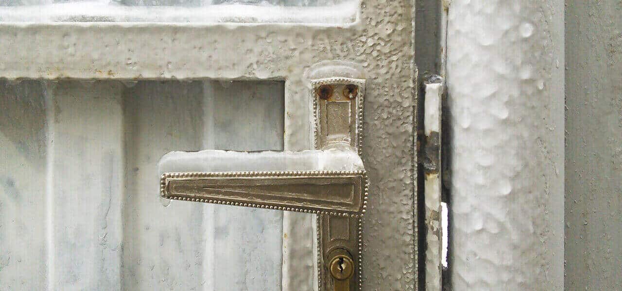 A metal door handle frozen over with a thick layer of ice is attached to a frosty door. The door and surrounding metal surfaces are covered in frost and ice, signaling the need for a skilled locksmith to navigate these extremely cold weather conditions.