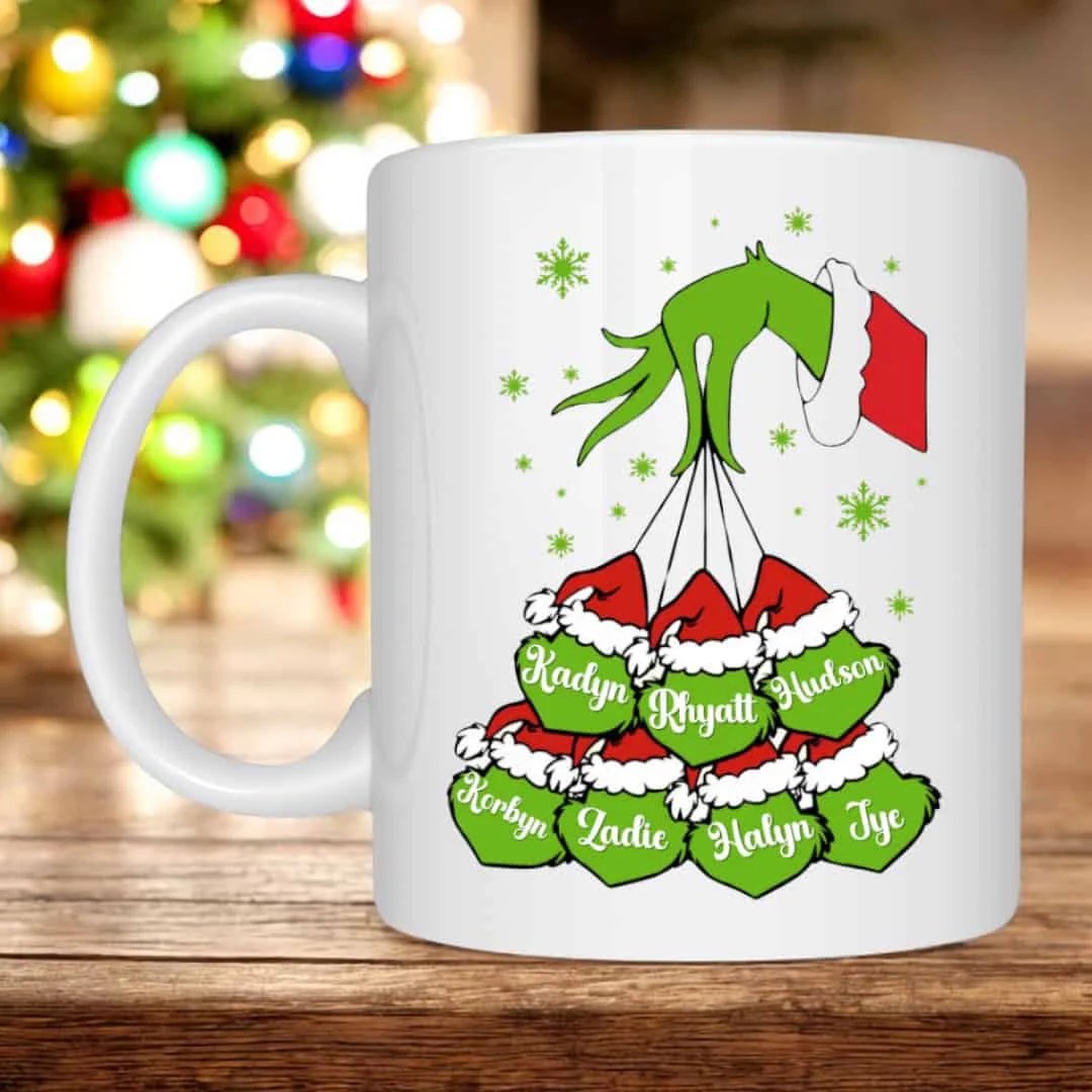 Christmas Gmones in Lights Classic T-Shirt 19 A Personalised Grinch Head Mug features a white Grinch face, green cartoon hand in a Santa sleeve holding up to 7 red and green Santa hats with custom names. A blurred Christmas tree is visible in the background.