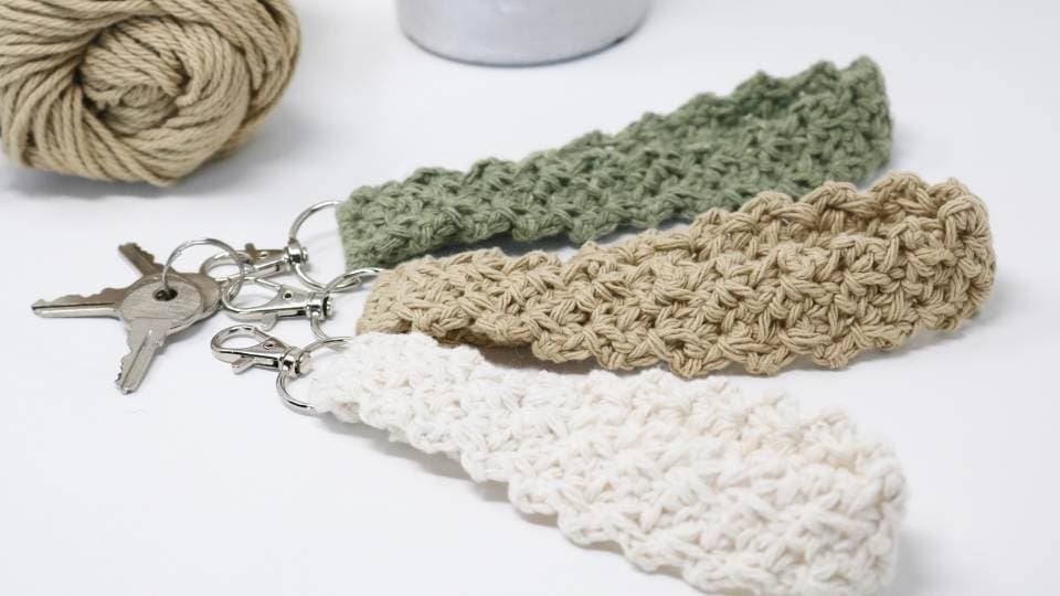 Three crocheted keychain wristlets in green, tan, and white lie on a white surface next to a ball of tan yarn and a set of keys—perfect for trying out a new free crochet pattern.