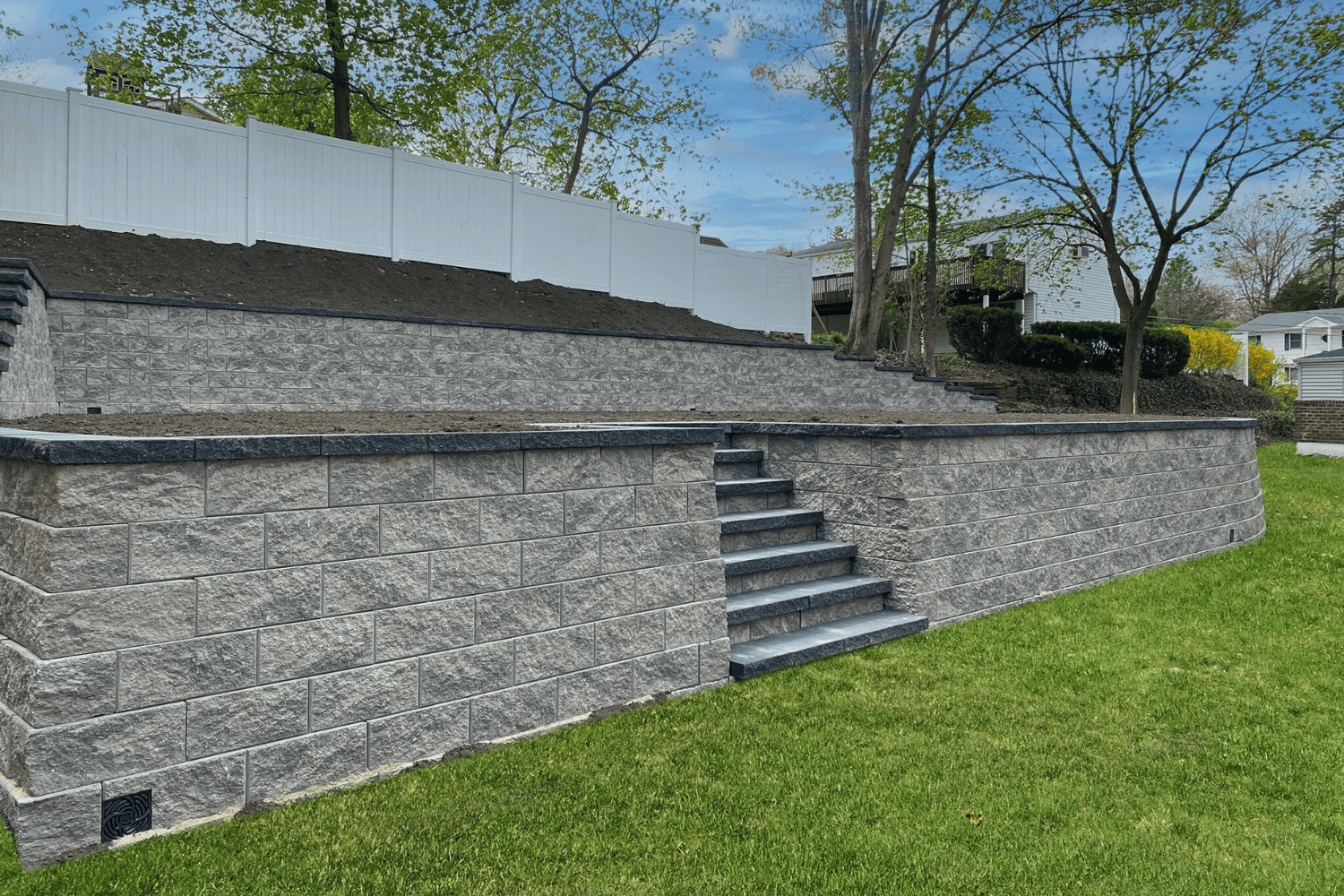 Stone retaining wall with steps leading up to a higher level of soil, bordered by white fencing and surrounded by grass and trees—an ideal example of how retaining walls add structure and beauty to your landscape.