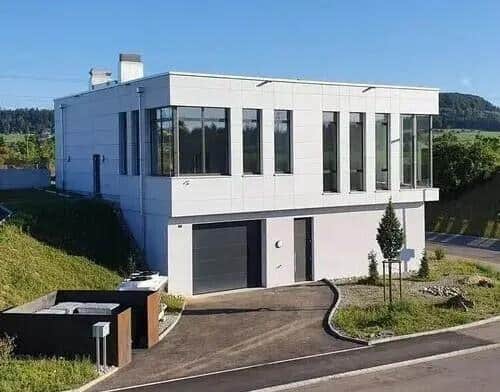 Modern white building with large windows and a dark gray garage door. A small, green tree stands in front of the building. The building is likely a European cryonics facility for body freezing, offering human preservation services.