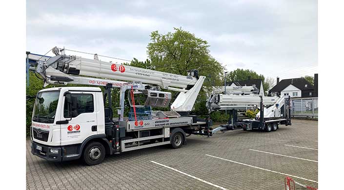 Lkw mit Hubarbeitsbühne steht auf einem gepflasterten Parkplatz, Baum im Hintergrund, Arbeitskorb ausgefahren, teilweise bewölkter Himmel.