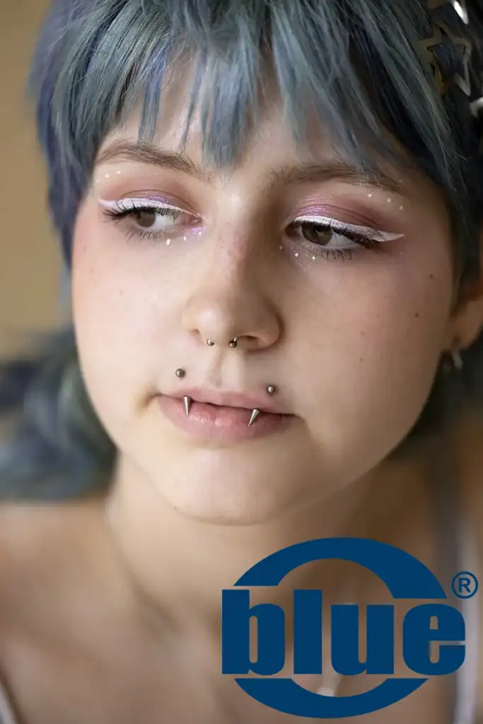 Close-up portrait of a person with blue hair and pink glittery eye makeup, white liner, freckles, and multiple nose and lip piercings; blue logo overlaid at the bottom right.