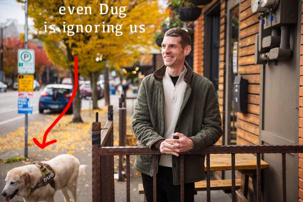 A man stands by a wooden railing on a city street. A dog wearing a vest labeled DUG walks on the sidewalk nearby. Text on the image reads even Dug is ignoring us with an arrow pointing to the dog.