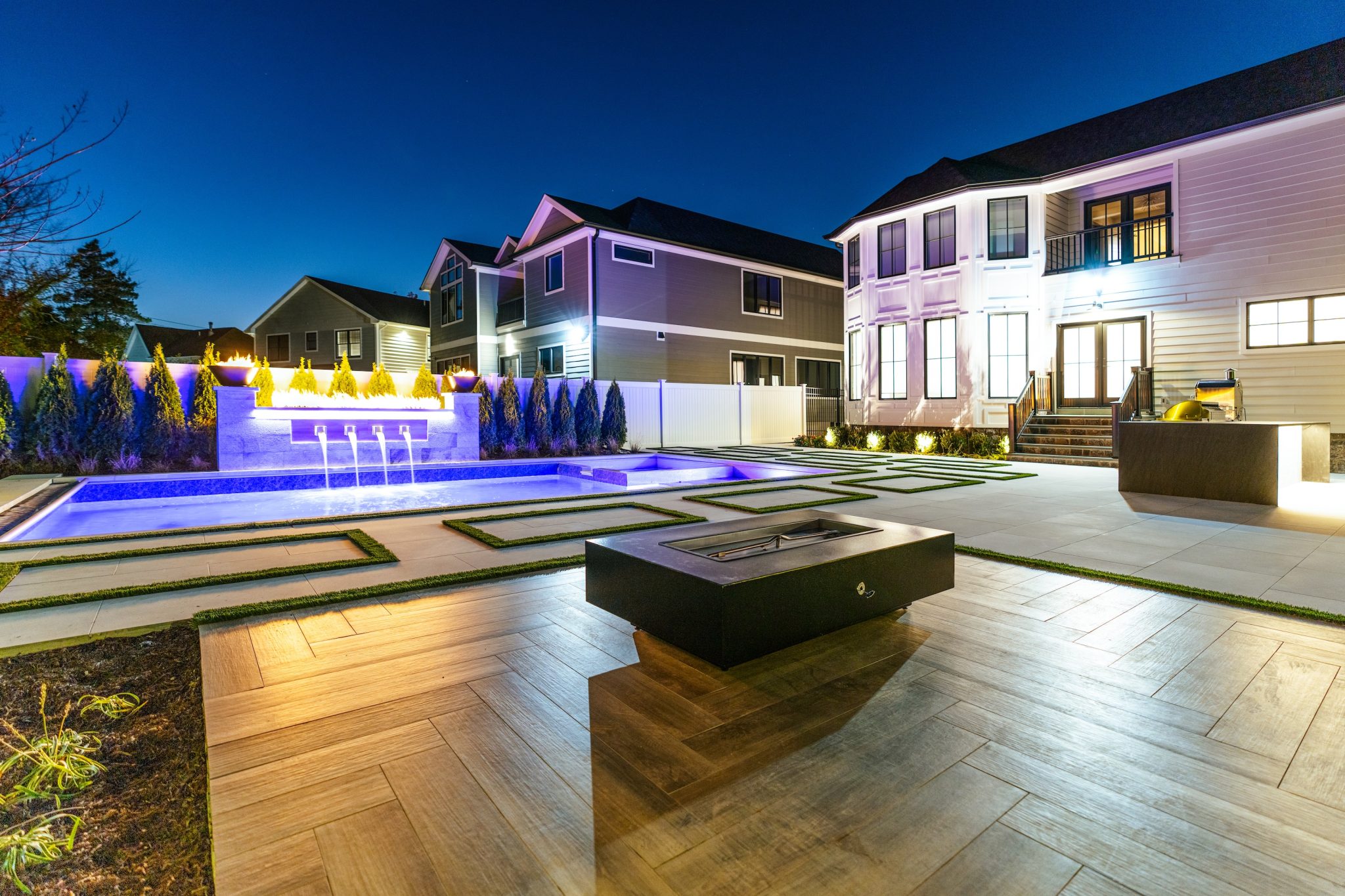 Contemporary backyard with illuminated pool, water features, manicured landscaping, a fire pit, and two modern houses at dusk.