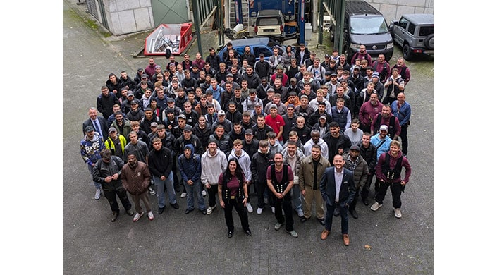 Bild von Begrüßung von Azubis mit ihren Lehrlingswarten Eine große Gruppe von Menschen steht draußen auf einer Straße und blickt in die Kamera. Im Hintergrund sind Autos und Gebäude zu sehen.