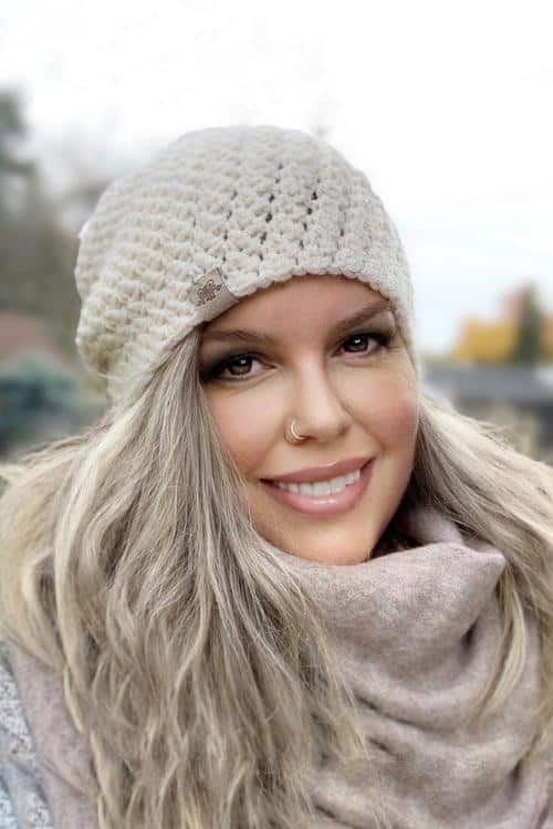 Smiling woman with long, wavy blond hair wears a beige beanie and matching scarf outside on a cloudy day. No text visible. easy and free crochet slouch hat pattern