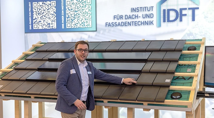 Ein Mann im Anzug präsentiert moderne Dach- und Fassadenplatten an einem Ausstellungsstand. Im Hintergrund ist ein Schild mit Text und QR-Code zu sehen.