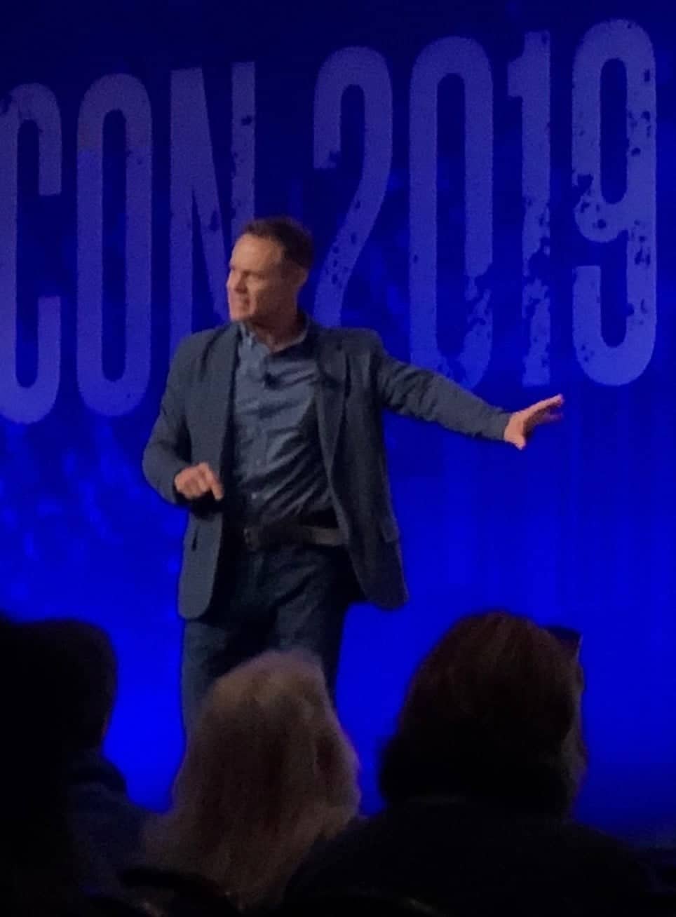 Paul Holes speaking on a stage in front of a large blue CrimeCon 2019 backdrop, gesturing with one hand.