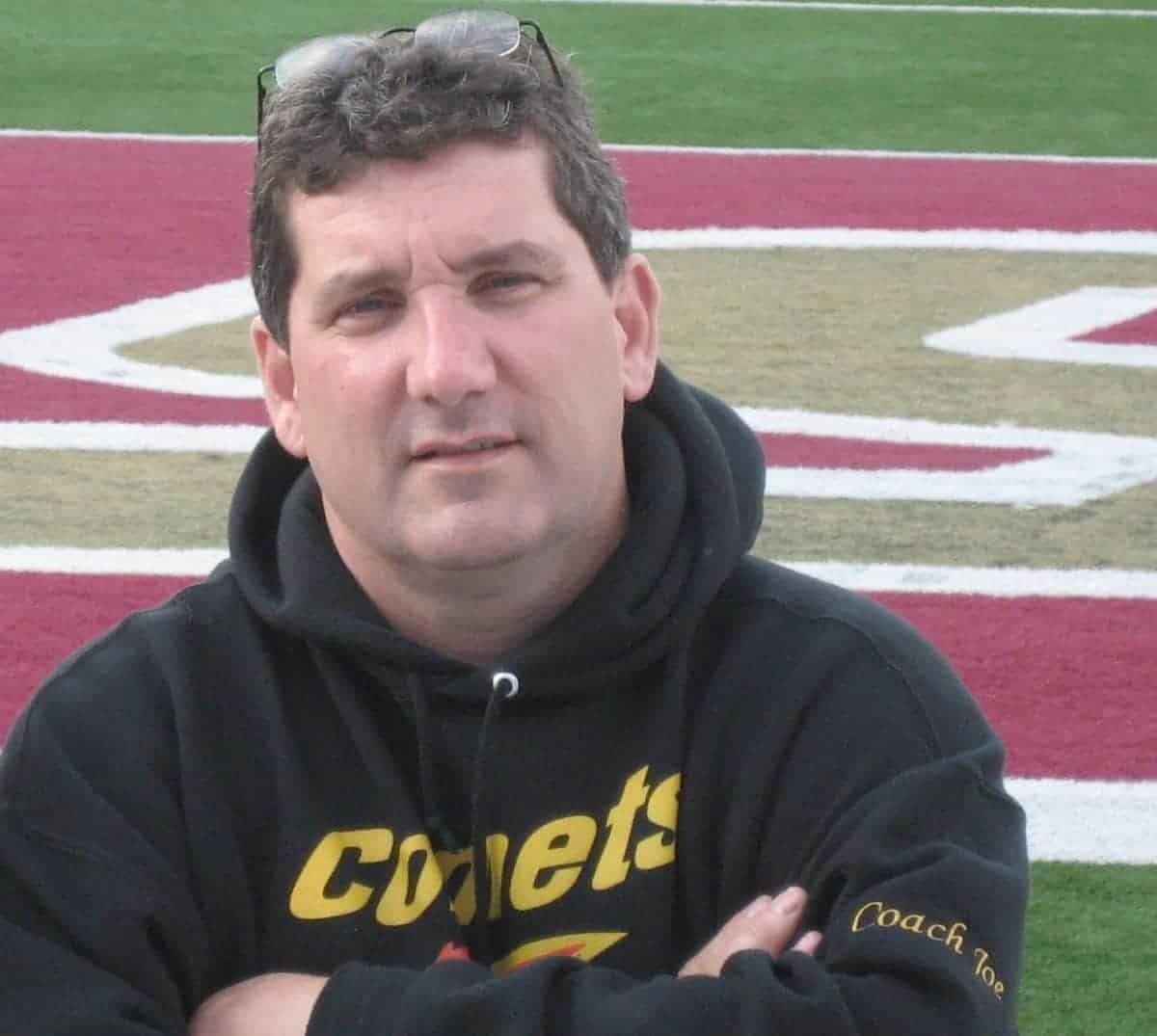 Coach Joe, wearing a black "Comets" hoodie, stands with arms crossed on a football field. He has glasses on top of his head. The background shows the field with red and green colors.