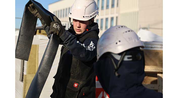 Person mit weißem Schutzhelm und dunkler Arbeitskleidung arbeitet im Freien, im Hintergrund ist ein modernes Gebäude zu sehen.