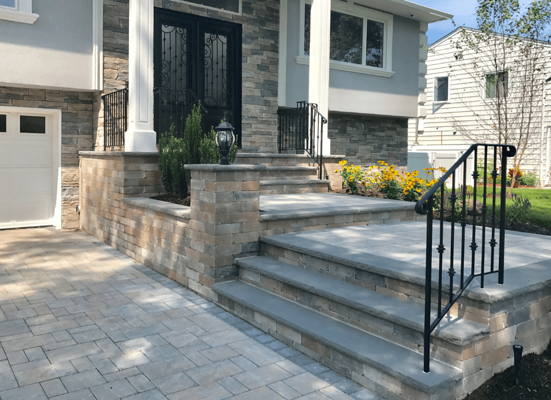 Stone stairs with metal railings lead to a black door on a brick facade house. Paved walkway and yellow flowers on the right.