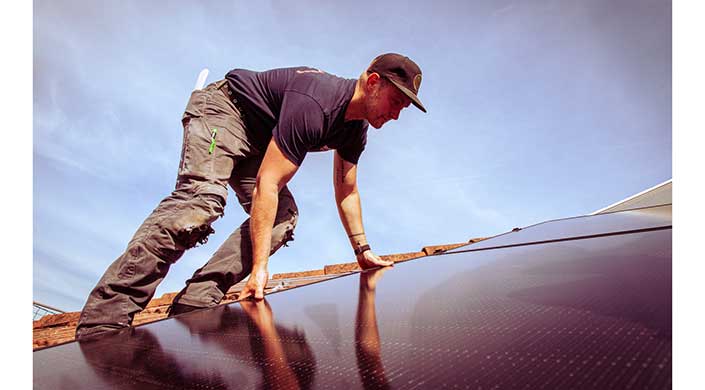 Ein Mann montiert tagsüber eine Solaranlage auf einem Dach unter klarem Himmel.