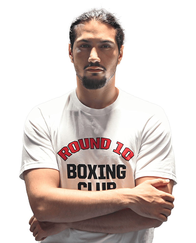 A confident man with sharp dark hair and a distinguished goatee stands with arms crossed at Round 10 Boxing Club in Al Quoz, Dubai. Clad in our signature white club t-shirt, he embodies the spirit of excellence and discipline that defines our gym—an ideal setting for those committed to mastering the art of boxing under expert guidance.