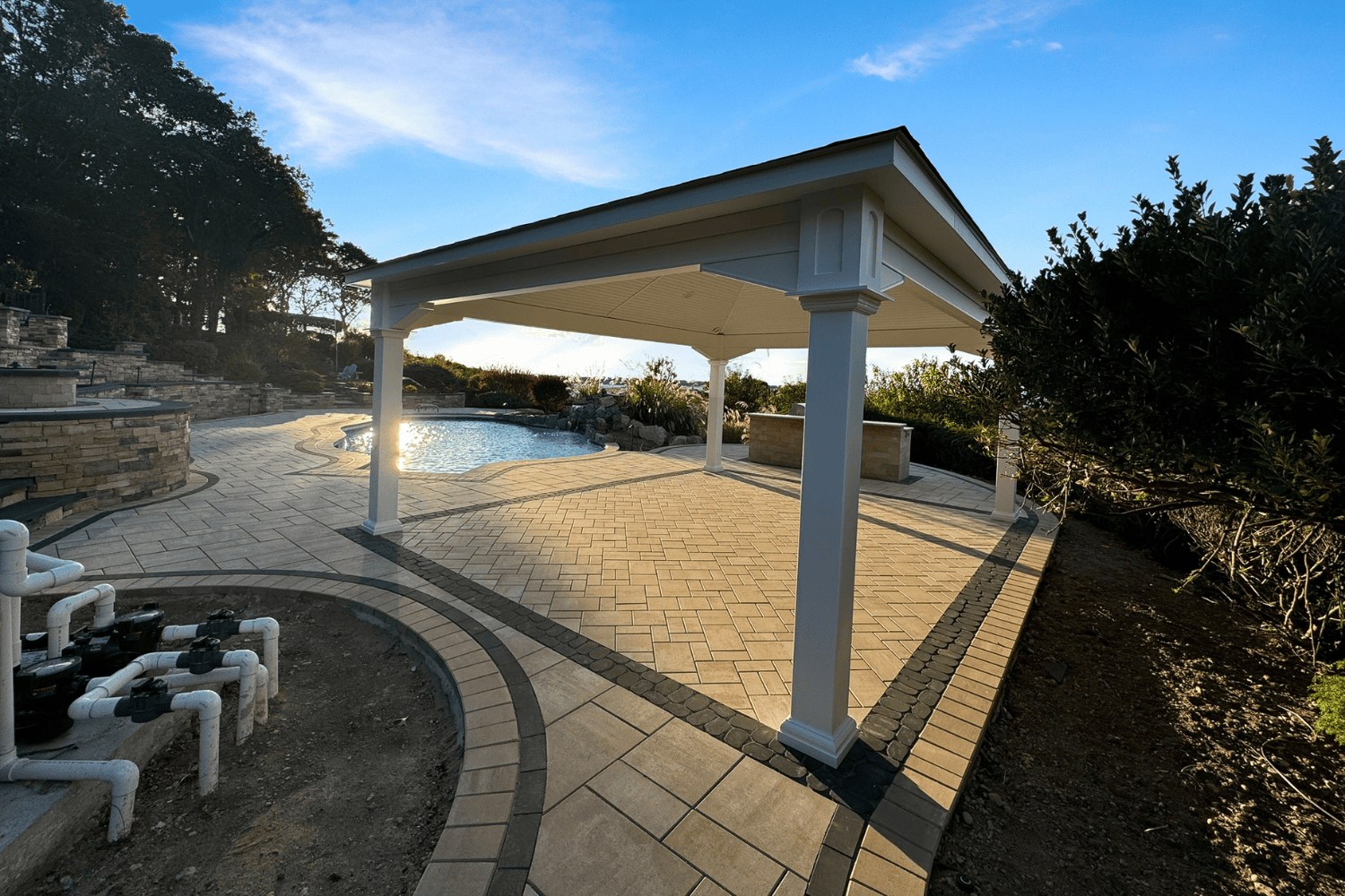 A covered patio with white columns and a paved surface overlooks a backyard pool, surrounded by landscaping, outdoor plumbing, and elegant pergolas for added shade.