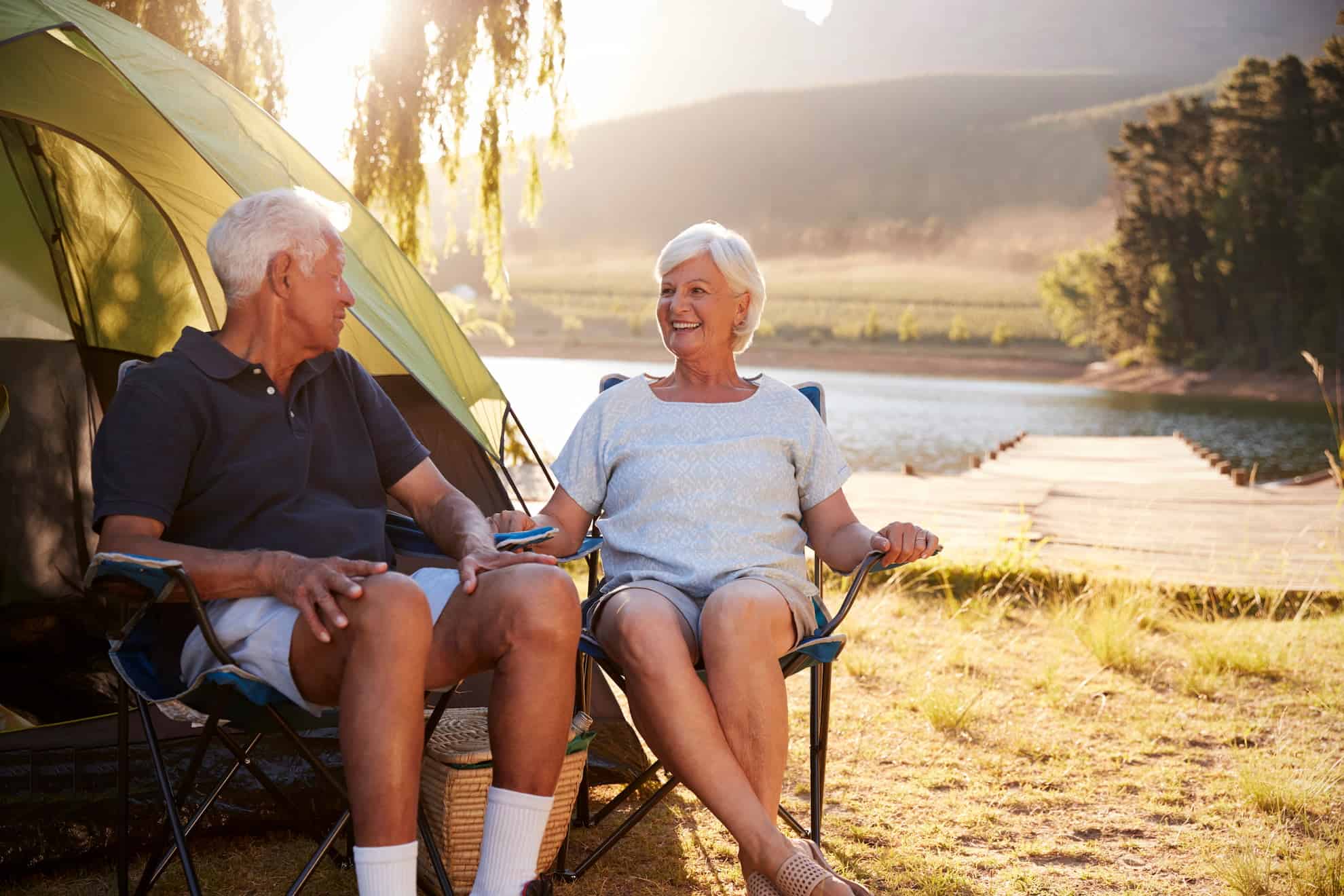 Smiling retired couple relaxing in camping chairs by a lake near a wooden dock, enjoying the outdoors together.