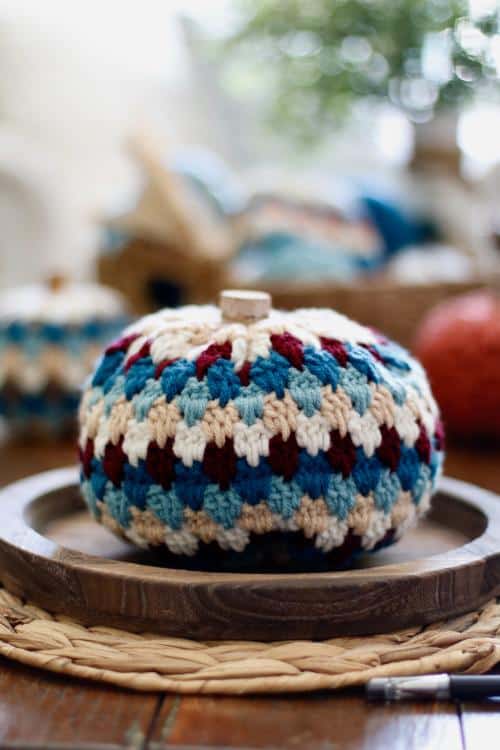 Multicolored crochet pumpkin with blue, burgundy, and beige patterns on a wooden plate atop a woven placemat; blurred background. Free crochet pattern