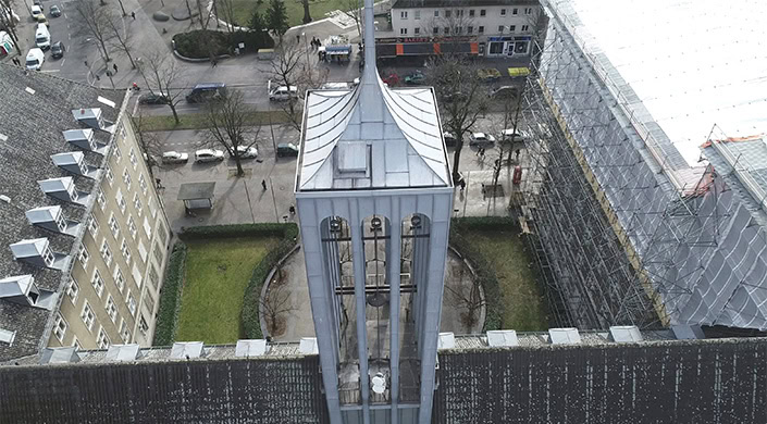 Ein moderner Glockenturm aus Metall mit spitzem Dach, Glocken sichtbar, im Hintergrund Bäume und Gebäude in einer Stadt.