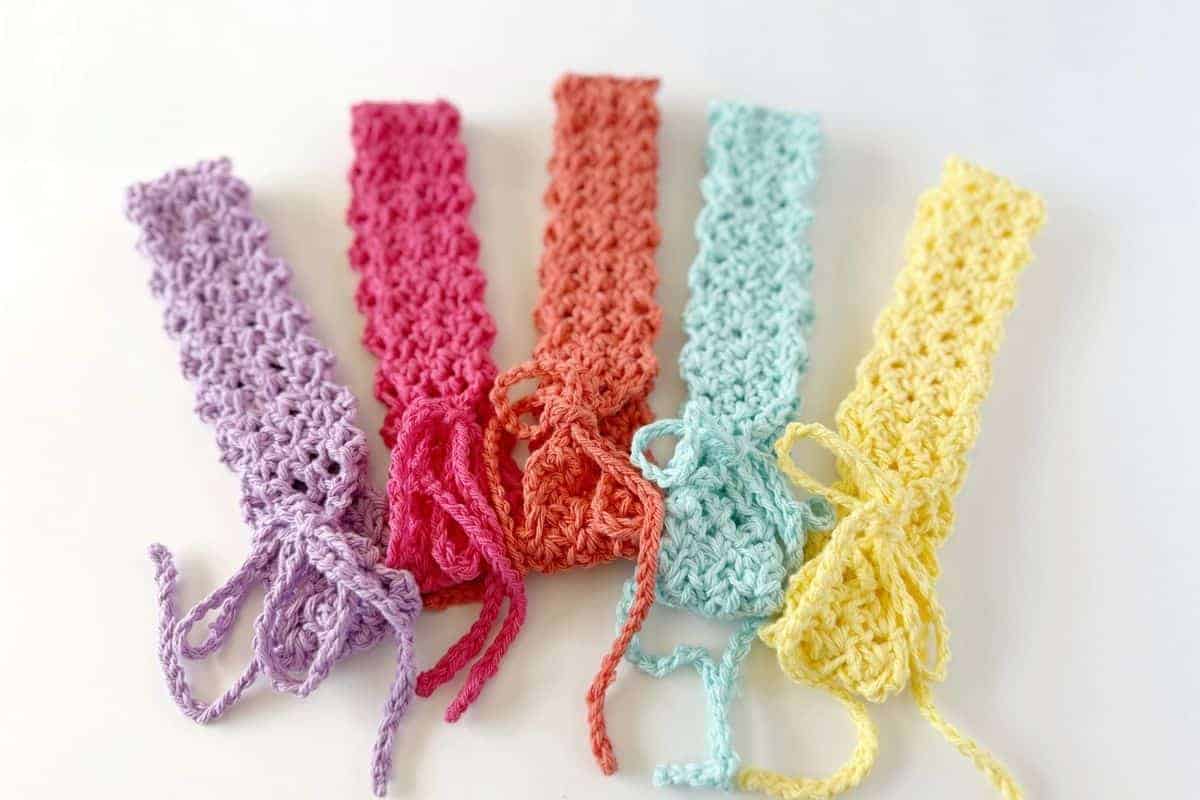 Five crocheted headbands in purple, pink, red, blue, and yellow lay side by side on a white background, each featuring a tied string at one end. No visible text or promotional content.