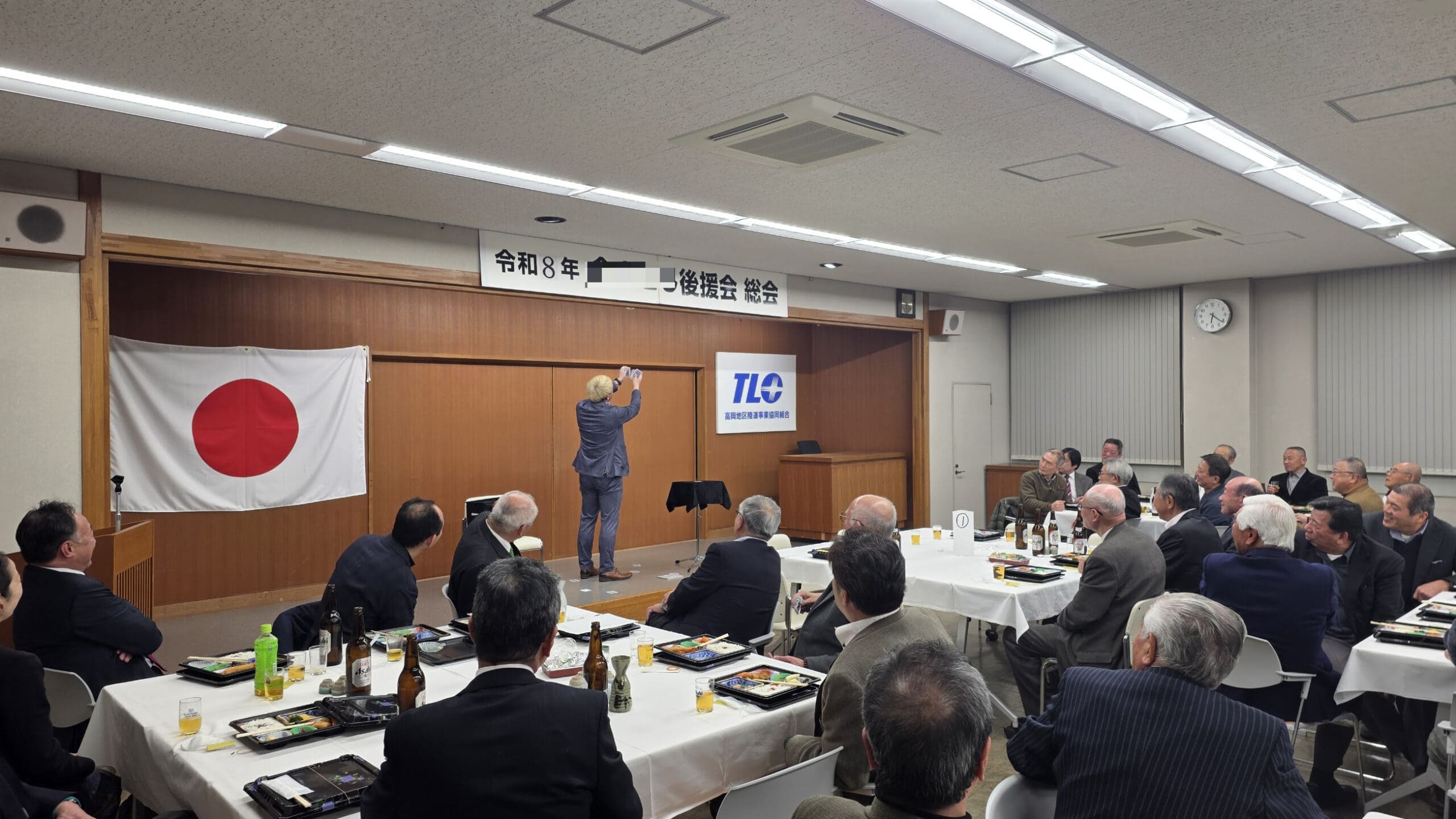 射水市テルシのパーティーでマジックショー! 3 ステージ上でマジックを披露するコンプレッサー。横断幕と日の丸が掲げられた会場