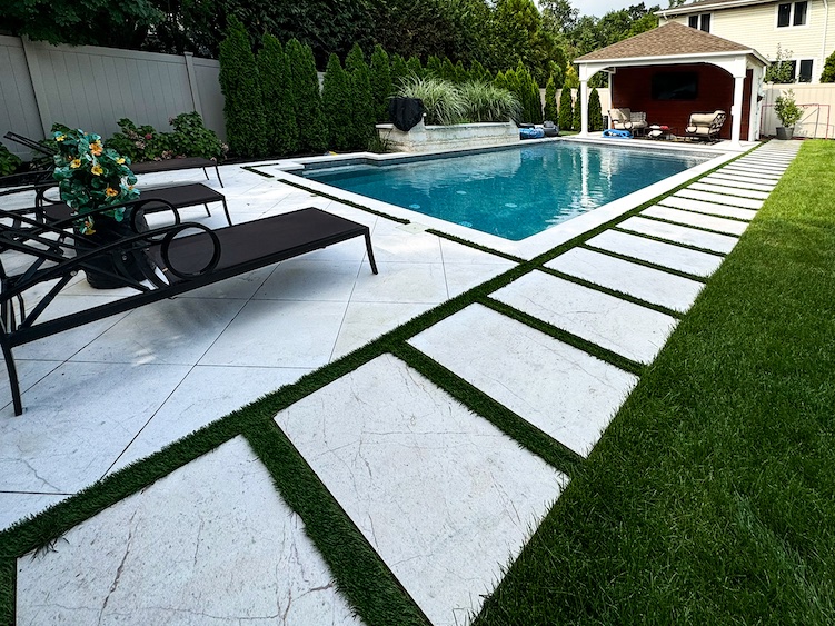 Rectangular swimming pool with chaise lounge chairs on a tiled deck, bordered by grass and shrubs, with a covered outdoor seating area in the background.