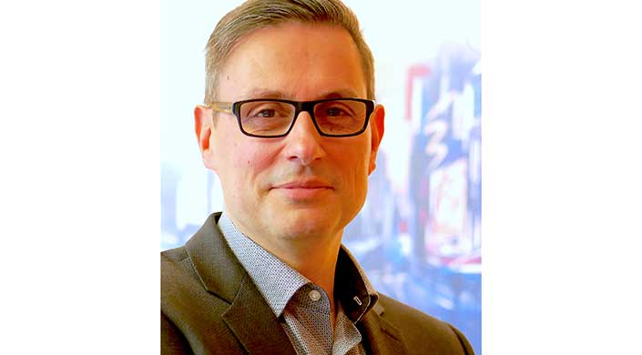 Mann mit Brille und Hemd blickt in die Kamera, Hintergrund unscharf.