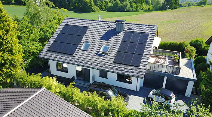 Modernes Haus mit schwarzen Dachziegeln, Solarpanelen und Oberlichtern, daneben parkt ein schwarzes Auto, im Hintergrund grüne Wiese und Bäume.