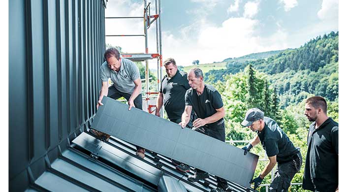 Mehrere Männer installieren Solarpaneele auf einem Dach, mit einem Gerüst im Hintergrund und einer ländlichen Umgebung.