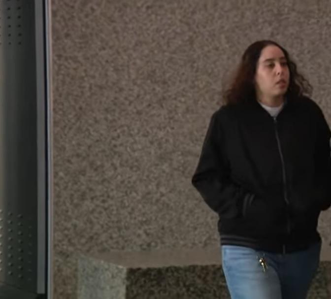A woman with long, brown hair, wearing a black jacket and light blue jeans, walks past a textured concrete wall. She is likely a former correctional officer facing sexual charges, connected to the Heather Mack prison abuse case.